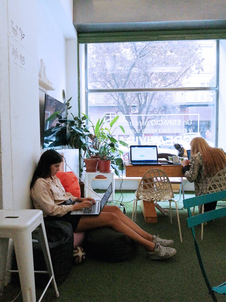 trabajar-en-coworking Dos emprendedoras salen cómodamente trabajando en el espacio de coworking Freeland, en Madrid, una de ellas con el ordenador en las rodillas, la otra en una mesa. Rodeadas por mobiliario colorido y plantas.